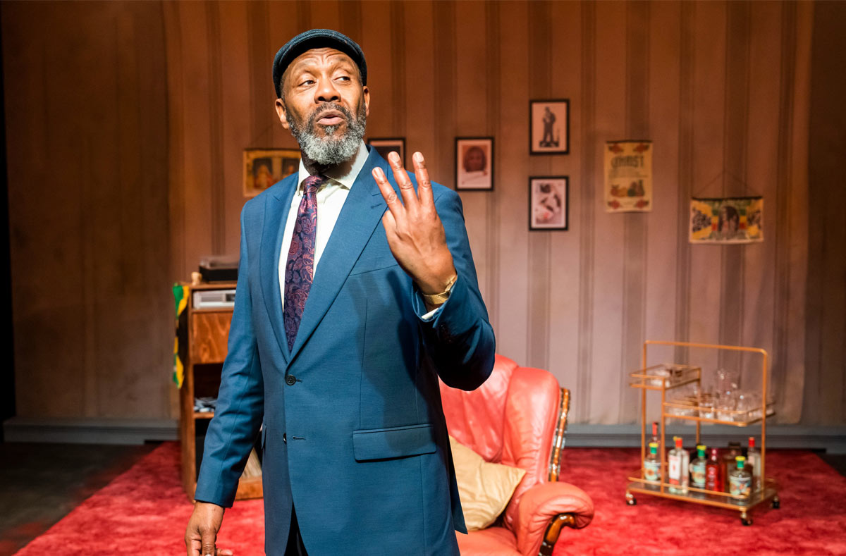 August is a black man in his sixties. He is wearing a blue suit, cap and purple tie. He is mid-speech, showing the audience three fingers. In the background you can see striped paper, a red armchair and carpet, a bar cart and family portraits on the wall.